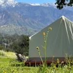 Canvas tent set up in a scenic green valley with wildflowers and snow-capped mountains in the background.