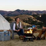 Couple roasting marshmallows over a campfire in front of a canvas tent with mountain views during a summer camping trip