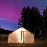 Canvas tent glowing under a starry night sky with purple and blue hues, surrounded by trees — perfect for stargazing