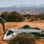 Camper setting up a cabin tent on a desert terrain with mountains in the background.