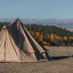 Camper securing tent pegs in a scenic mountain campsite with fall foliage and canvas tent