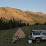 Summer camping scene with a couple by a campfire, canvas tent, and truck in a scenic mountain valley at sunset