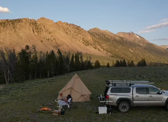 Summer camping scene with a couple by a campfire, canvas tent, and truck in a scenic mountain valley at sunset