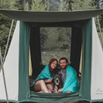 Couple with dog relaxing in a White Duck cabin tent, wrapped in blankets, enjoying a cool and comfortable camping sleep in the forest.