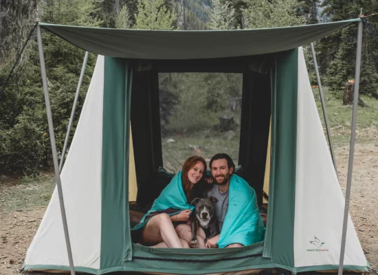 Couple with dog relaxing in a White Duck cabin tent, wrapped in blankets, enjoying a cool and comfortable camping sleep in the forest.