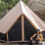 Person camping with their dog in front of a canvas tent surrounded by rocks and trees.