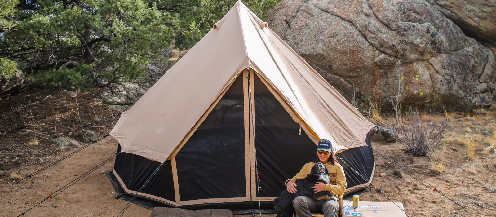 Person camping with their dog in front of a canvas tent surrounded by rocks and trees.