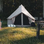 Two women enjoying a conversation outside a canvas bell tent during a camping trip, with gear and picnic setup nearby.
