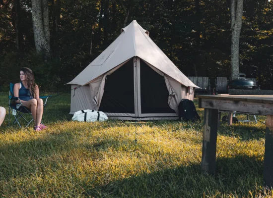 Two women enjoying a conversation outside a canvas bell tent during a camping trip, with gear and picnic setup nearby.