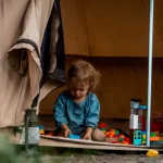 A young child playing with colorful building blocks at the entrance of a canvas camping tent, surrounded by nature.