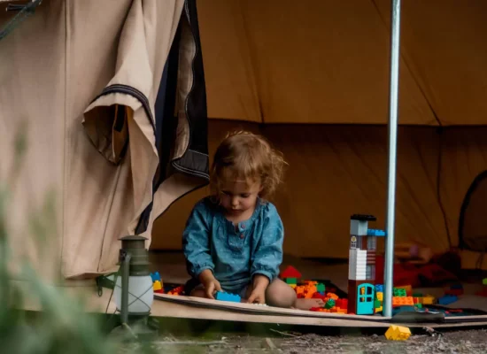 A young child playing with colorful building blocks at the entrance of a canvas camping tent, surrounded by nature.
