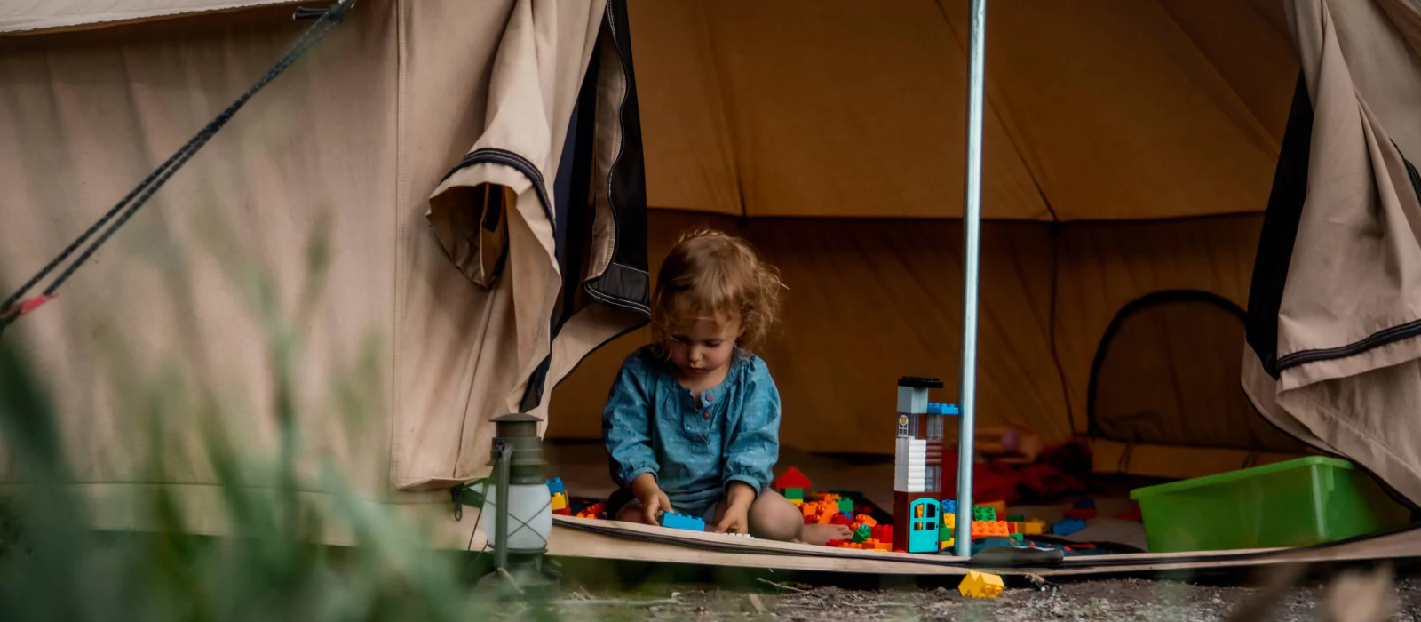 A young child playing with colorful building blocks at the entrance of a canvas camping tent, surrounded by nature.