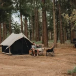 Man relaxing outside a canvas tent in a shaded forest campsite, demonstrating how to stay cool while camping in hot weather without A/C