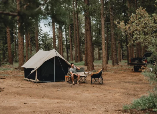 Man relaxing outside a canvas tent in a shaded forest campsite, demonstrating how to stay cool while camping in hot weather without A/C