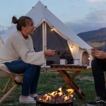 Couple camping by a cozy campfire outside a canvas tent, enjoying drinks and playing cards in nature