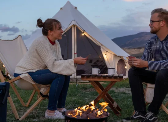 Couple camping by a cozy campfire outside a canvas tent, enjoying drinks and playing cards in nature