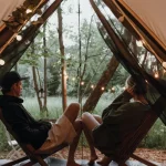 Couple relaxing inside a glamping tent decorated with string lights, enjoying a peaceful forest view and cozy outdoor escape