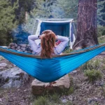 Person relaxing in a blue hammock between trees at a forest campsite, embracing nature for wellness and forest bathing benefits.