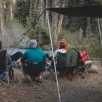 Group of friends relaxing in camping chairs near a lake, with a dog and a tent nearby, enjoying peaceful time by the water in the forest.