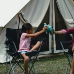 Mother and daughter clinking water bottles outside a canvas tent, promoting hydration and health during summer camping