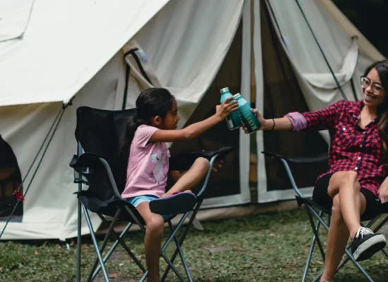 Mother and daughter clinking water bottles outside a canvas tent, promoting hydration and health during summer camping