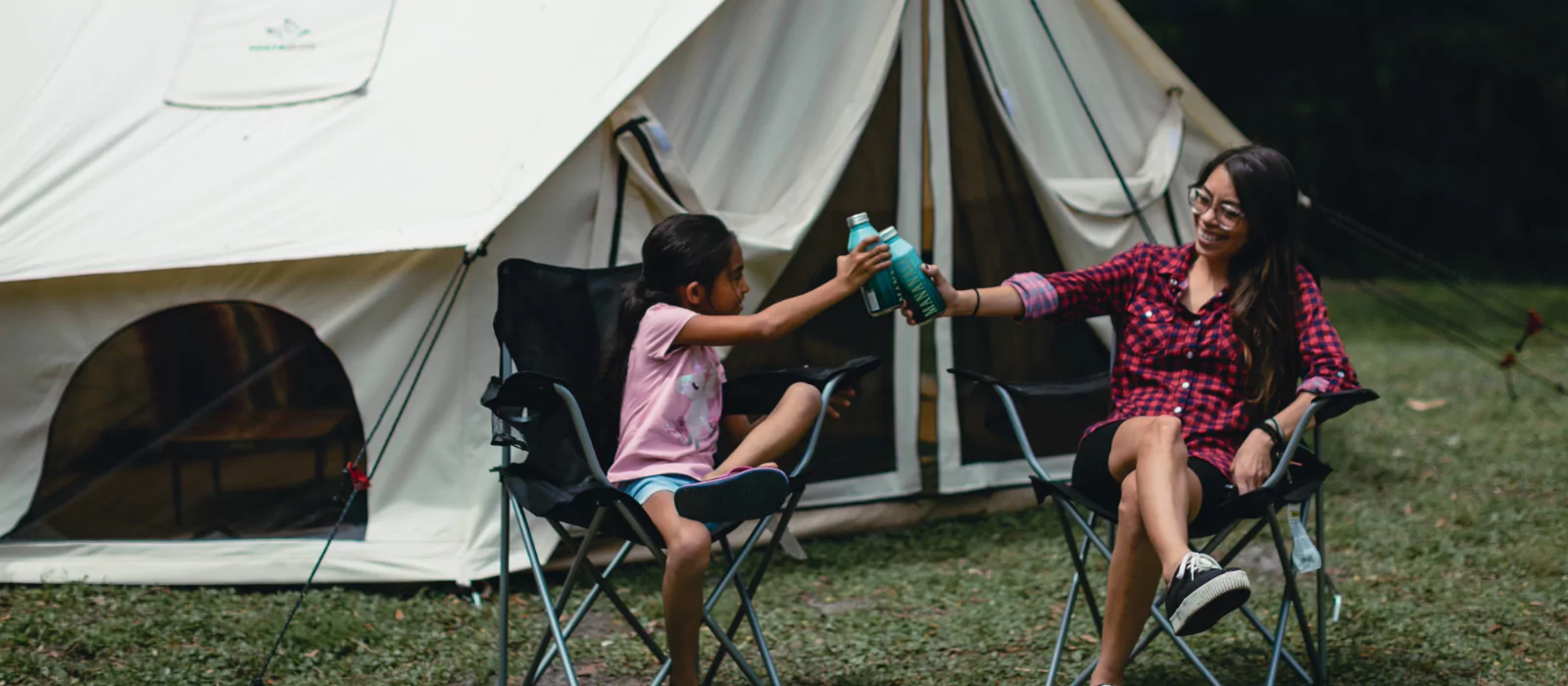 Mother and daughter clinking water bottles outside a canvas tent, promoting hydration and health during summer camping