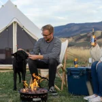Couple enjoying outdoor activities while camping in the mountains with their dog and a campfire outside a canvas tent.