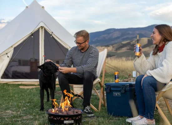 Couple enjoying outdoor activities while camping in the mountains with their dog and a campfire outside a canvas tent.