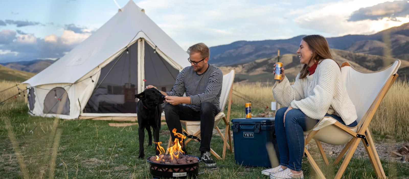 Couple enjoying outdoor activities while camping in the mountains with their dog and a campfire outside a canvas tent.