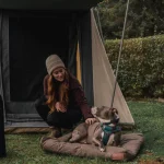 Woman outdoors with a dog, beside a canvas tent and White Duck canvas gear bag in a forested campsite setting.