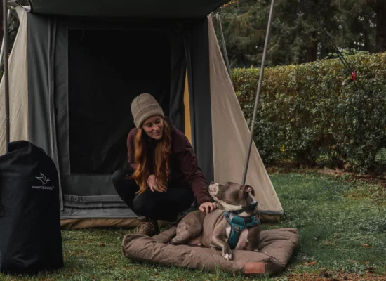 Woman outdoors with a dog, beside a canvas tent and White Duck canvas gear bag in a forested campsite setting.