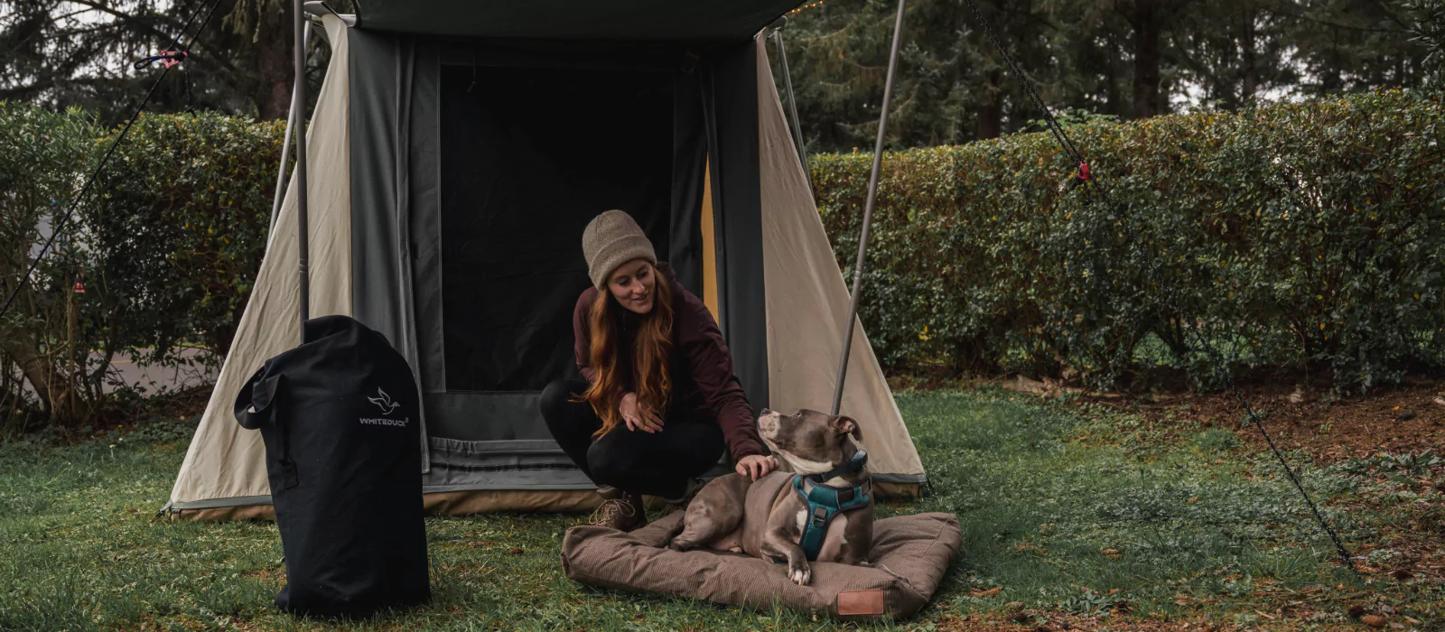 Woman outdoors with a dog, beside a canvas tent and White Duck canvas gear bag in a forested campsite setting.