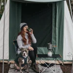 Woman enjoying a hot drink outside a White Duck cabin tent with her dog and a camping stove setup—perfect setting for easy summer camping meals.