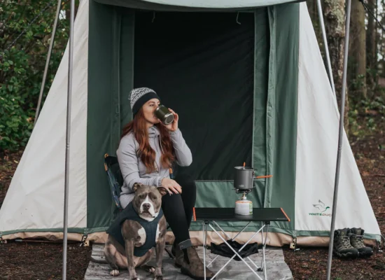 Woman enjoying a hot drink outside a White Duck cabin tent with her dog and a camping stove setup—perfect setting for easy summer camping meals.
