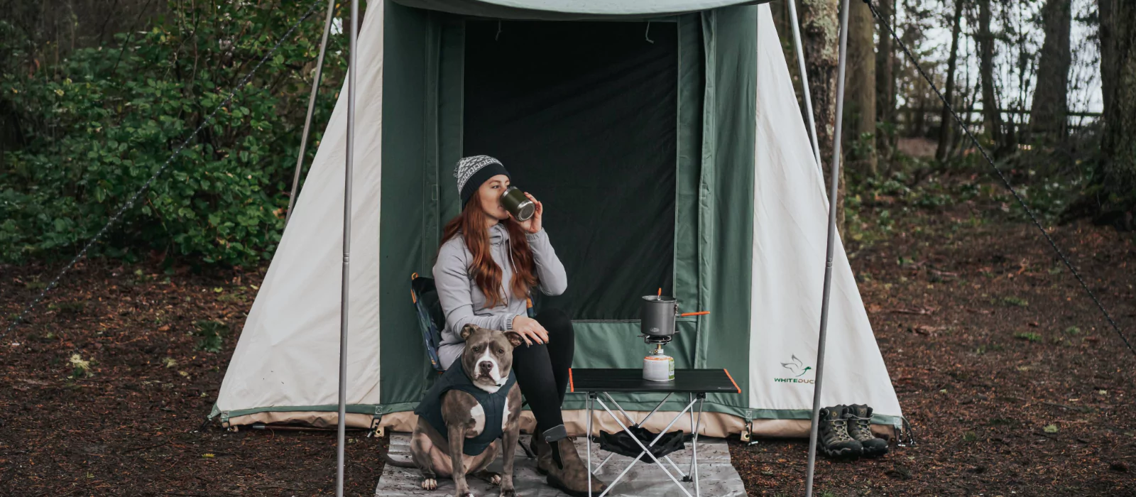 Woman enjoying a hot drink outside a White Duck cabin tent with her dog and a camping stove setup—perfect setting for easy summer camping meals.
