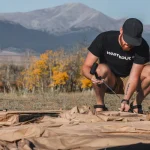 Man staking a canvas tent on a mountain campsite with fall foliage in the background