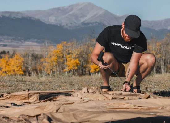Man staking a canvas tent on a mountain campsite with fall foliage in the background
