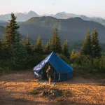 Blue bell tent set up on a scenic mountain campsite surrounded by pine trees at sunrise or sunset