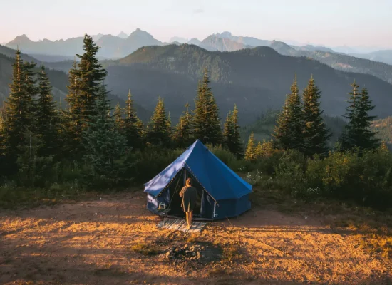 Blue bell tent set up on a scenic mountain campsite surrounded by pine trees at sunrise or sunset