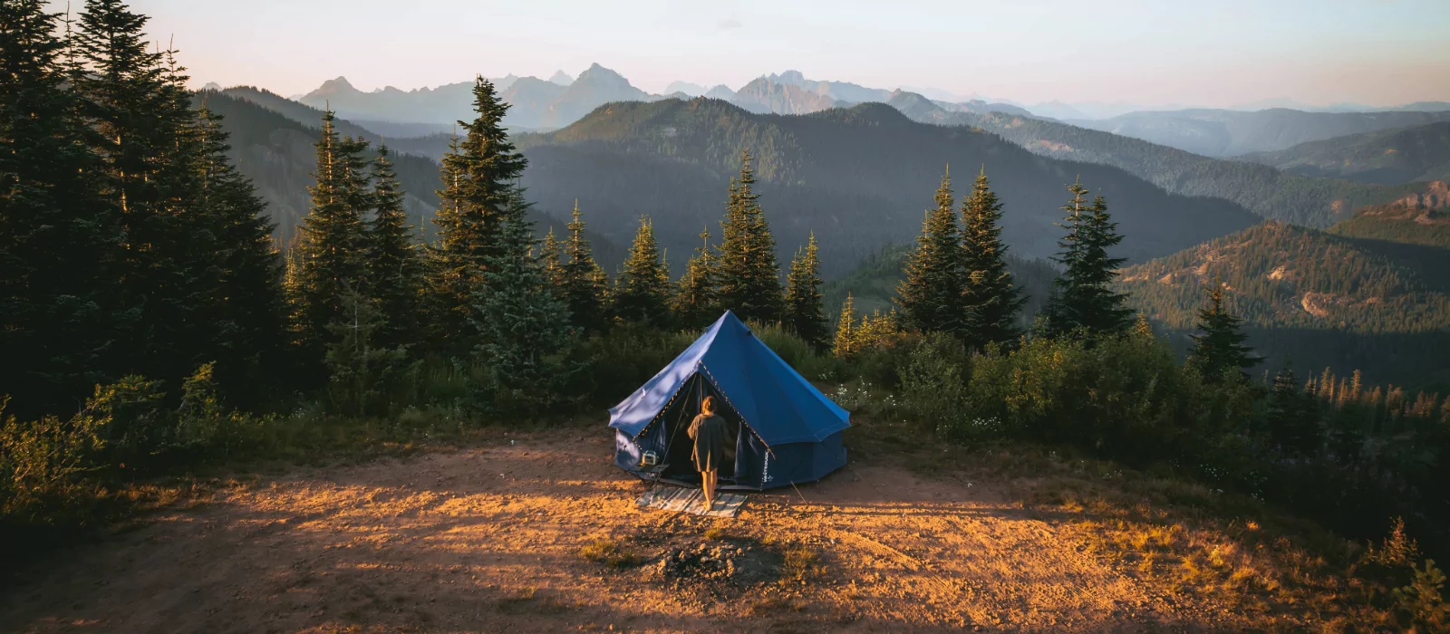 Blue bell tent set up on a scenic mountain campsite surrounded by pine trees at sunrise or sunset