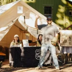 Group of people at a camping event standing near a canvas tent used as a merch booth