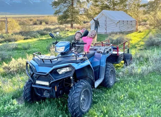 Two kids on an ATV during a camping scavenger hunt near a wall tent in a grassy field with mountain views.