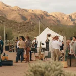 A decorated desert campsite with string lights, people socializing, and a canvas tent set against a backdrop of rugged mountains at sunset.
