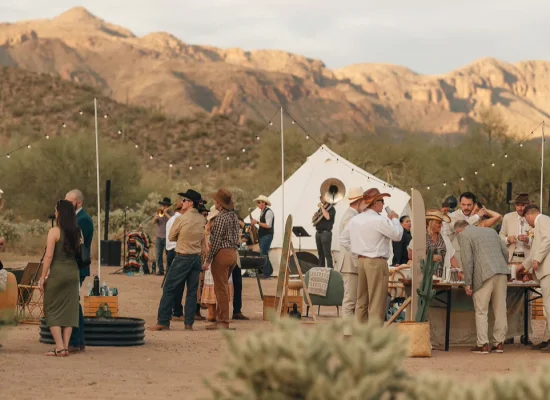 A decorated desert campsite with string lights, people socializing, and a canvas tent set against a backdrop of rugged mountains at sunset.