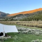 A white bell tent set up on a grassy field with mountains and pine trees in the background during a clear fall evening.