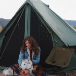 Woman solo camping with a husky dog in front of a canvas tent, surrounded by forest and mountains.