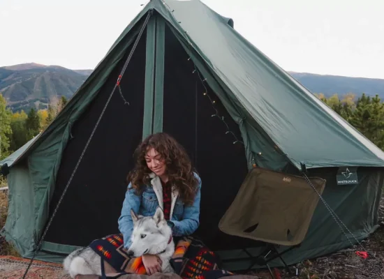 Woman solo camping with a husky dog in front of a canvas tent, surrounded by forest and mountains.