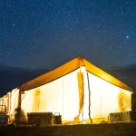 Glowing wall tent lit from inside under a clear starry night sky, surrounded by trees and camping gear