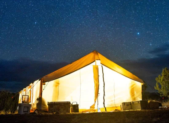 Glowing wall tent lit from inside under a clear starry night sky, surrounded by trees and camping gear