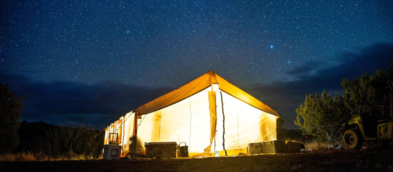 Glowing wall tent lit from inside under a clear starry night sky, surrounded by trees and camping gear